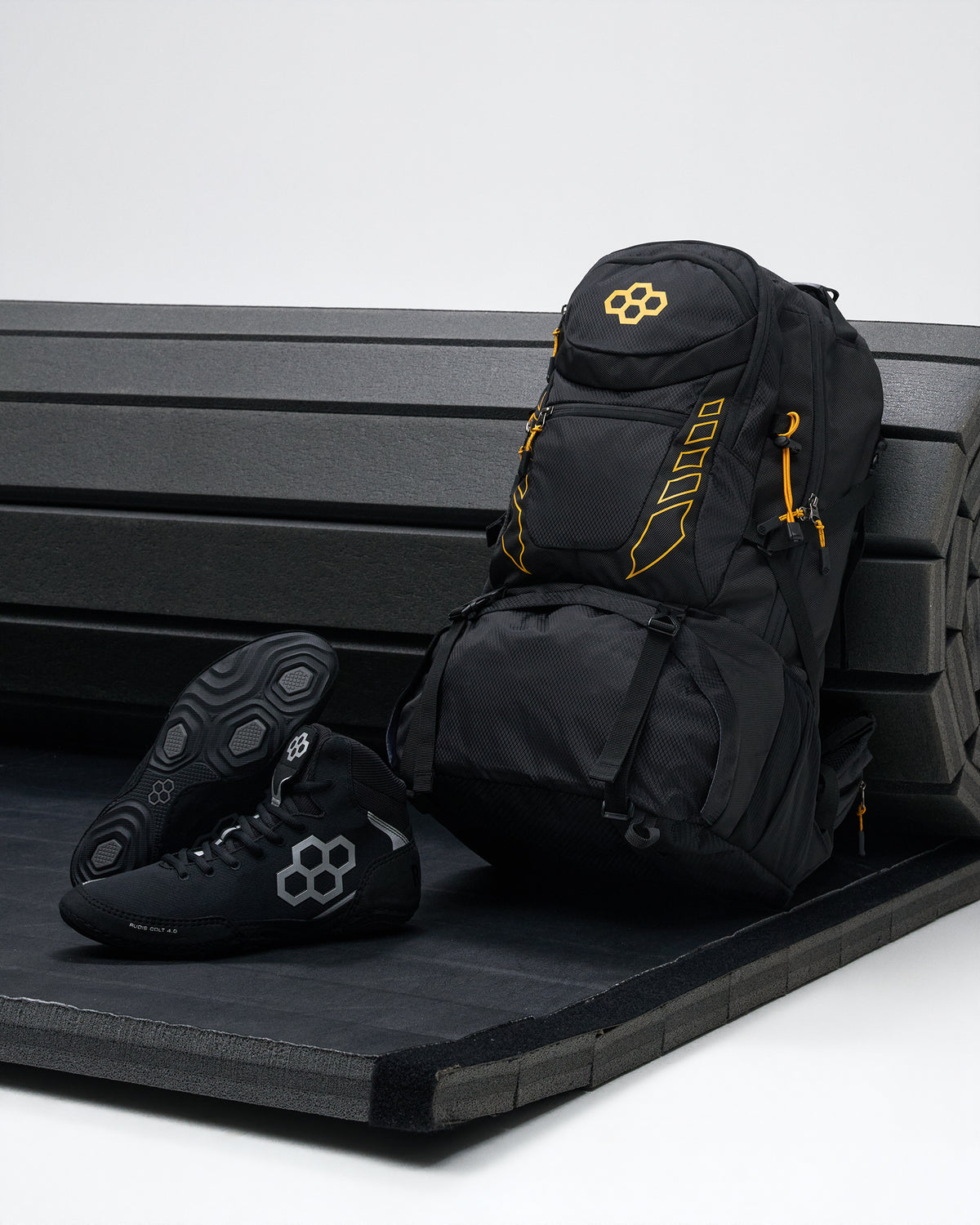 A black backpack with bright yellow accents and a matching black wrestling shoe with a gray geometric logo, displayed on a black bench against a light gray background.