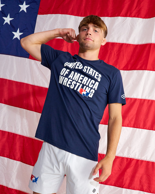 A man wearing a navy blue t-shirt with 'UNITED STATES OF AMERICA WRESTLING' printed in white and a small red, white, and blue logo, paired with white wrestling shorts, posed in front of a large American flag.