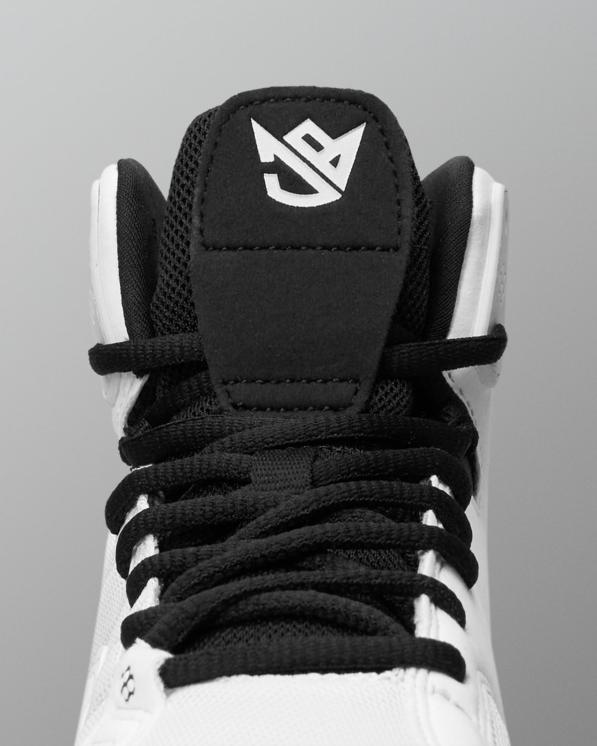 Close-up of a high-top wrestling shoe showing the black tongue patch with a white stylized logo, black laces, and white upper against a gray background.