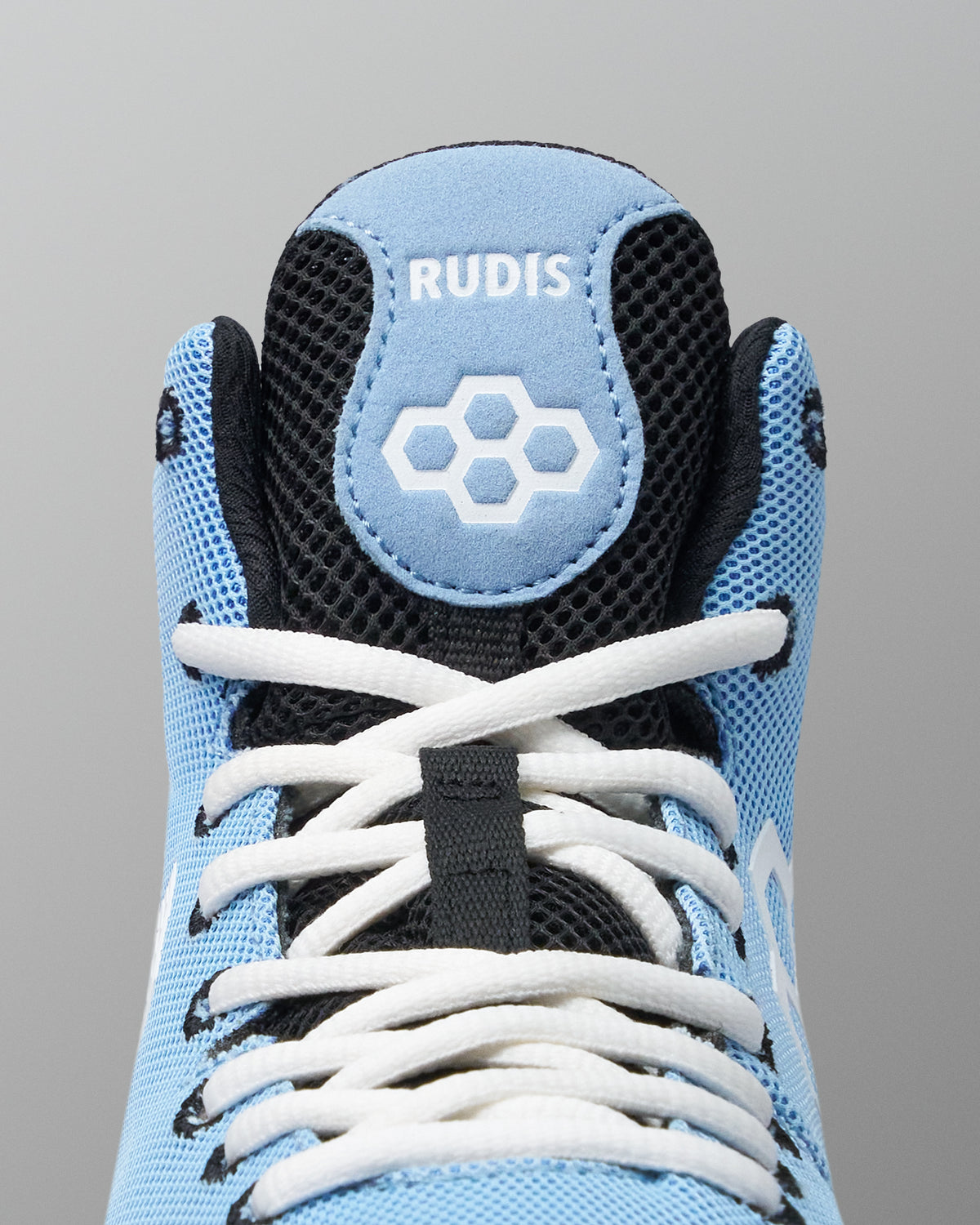 Close-up of the top of a light blue and black wrestling shoe with white laces. The tongue shows the brand name 'RUDIS' and a white geometric logo.
