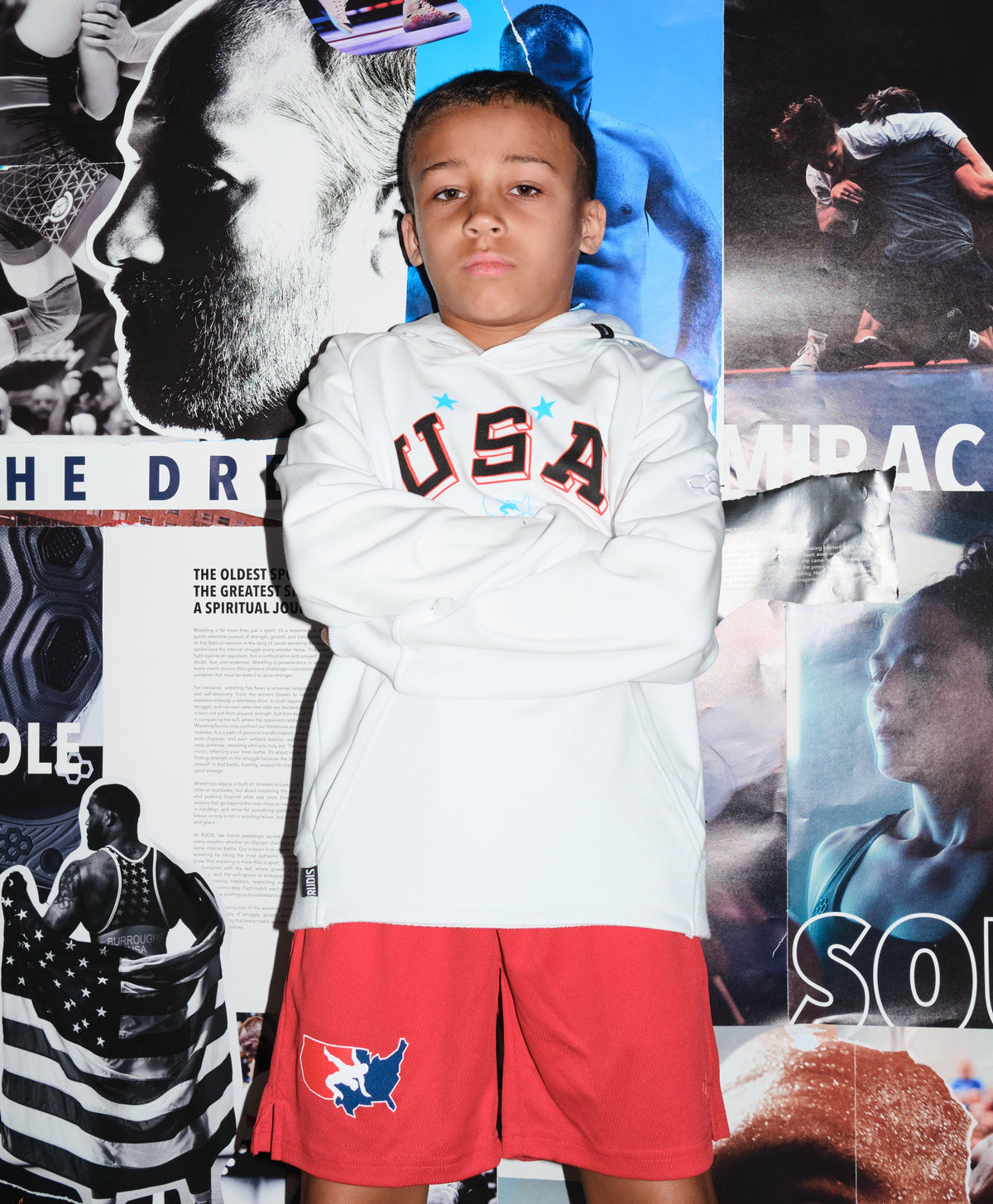 Beacon Burroughs, youth wrestler and son of Olympic champion Jordan Burroughs, wearing a white USA Wrestling hoodie and red USA Wrestling shorts by RUDIS, standing confidently in front of a wrestling mural — representing the next generation of wrestling.