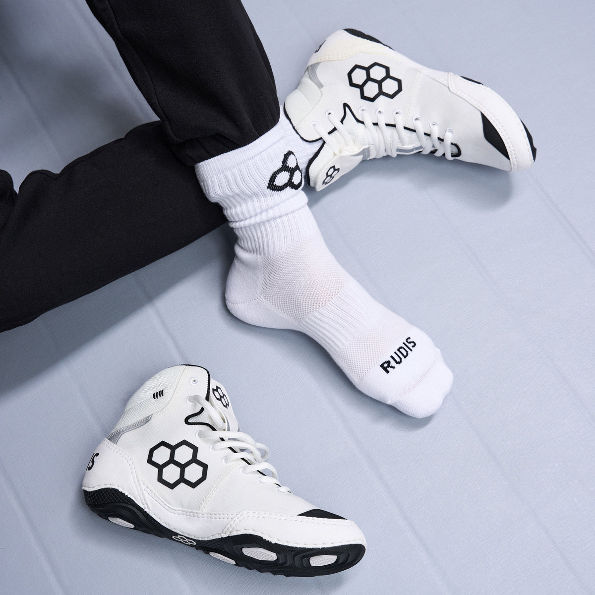 White wrestling shoes with black RUDIS Logo accents and a logo worn with white socks on a light gray floor.
