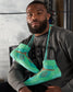 A man wearing a dark sweatshirt with LIBERTY text poses with vibrant green athletic shoes hanging around his neck showcasing their unique design and colors