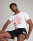 A male model poses confidently in a casual outfit consisting of a white t-shirt with a bold red wrestling graphic and black athletic shorts seated on a wooden stool against a light grey background