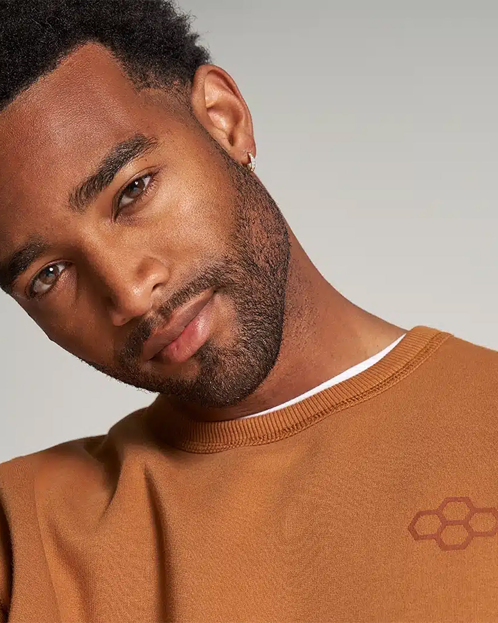 A man with a stylish look is posing in a brown sweatshirt showcasing his confident demeanor and contemporary sense of fashion