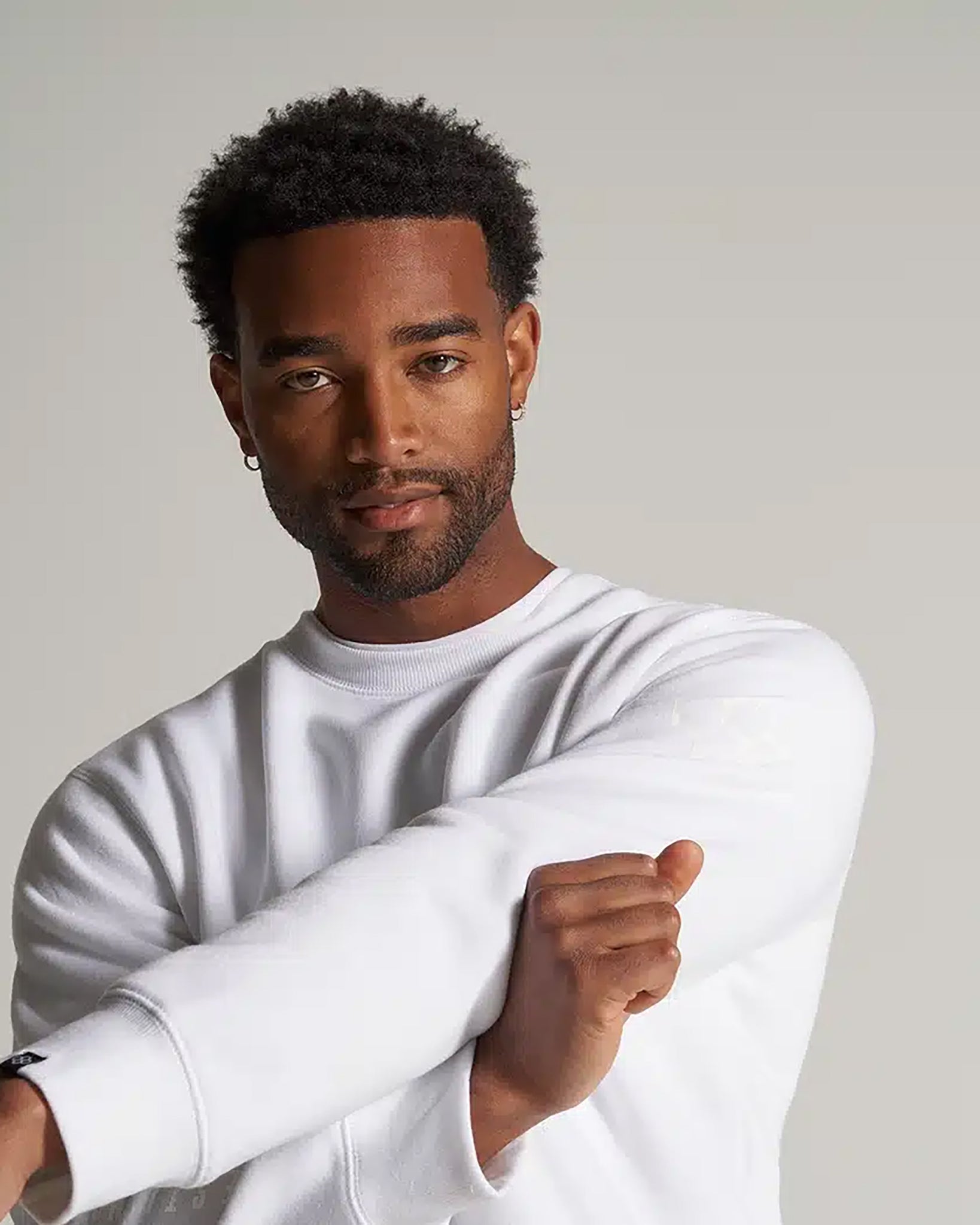 A man with short curly hair wearing a white sweatshirt with a relaxed expression showing one arm crossed over the other