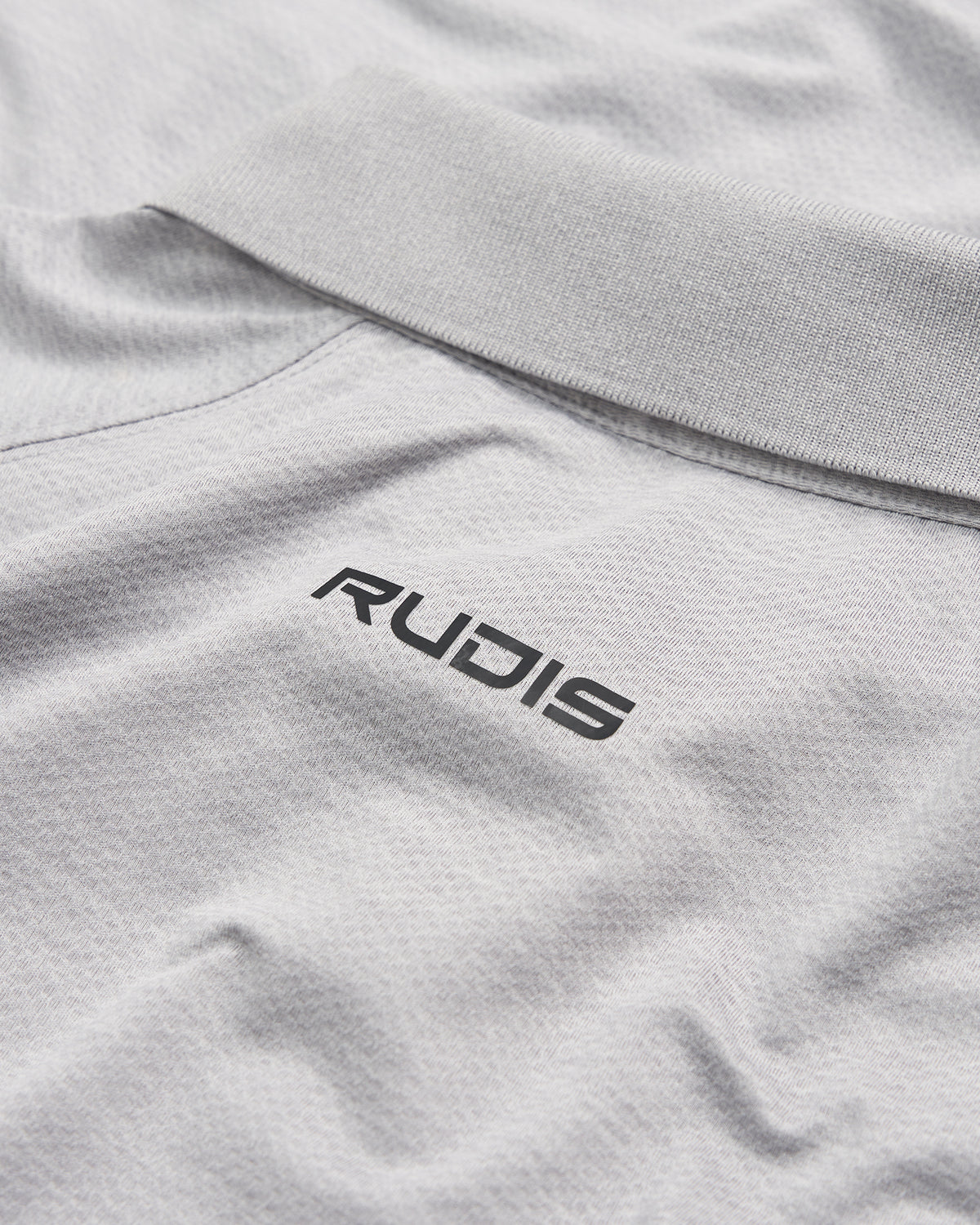 A close-up view of a light gray polo shirt featuring a black RUDIS logo on the collar area emphasizing a stylish and sporty aesthetic