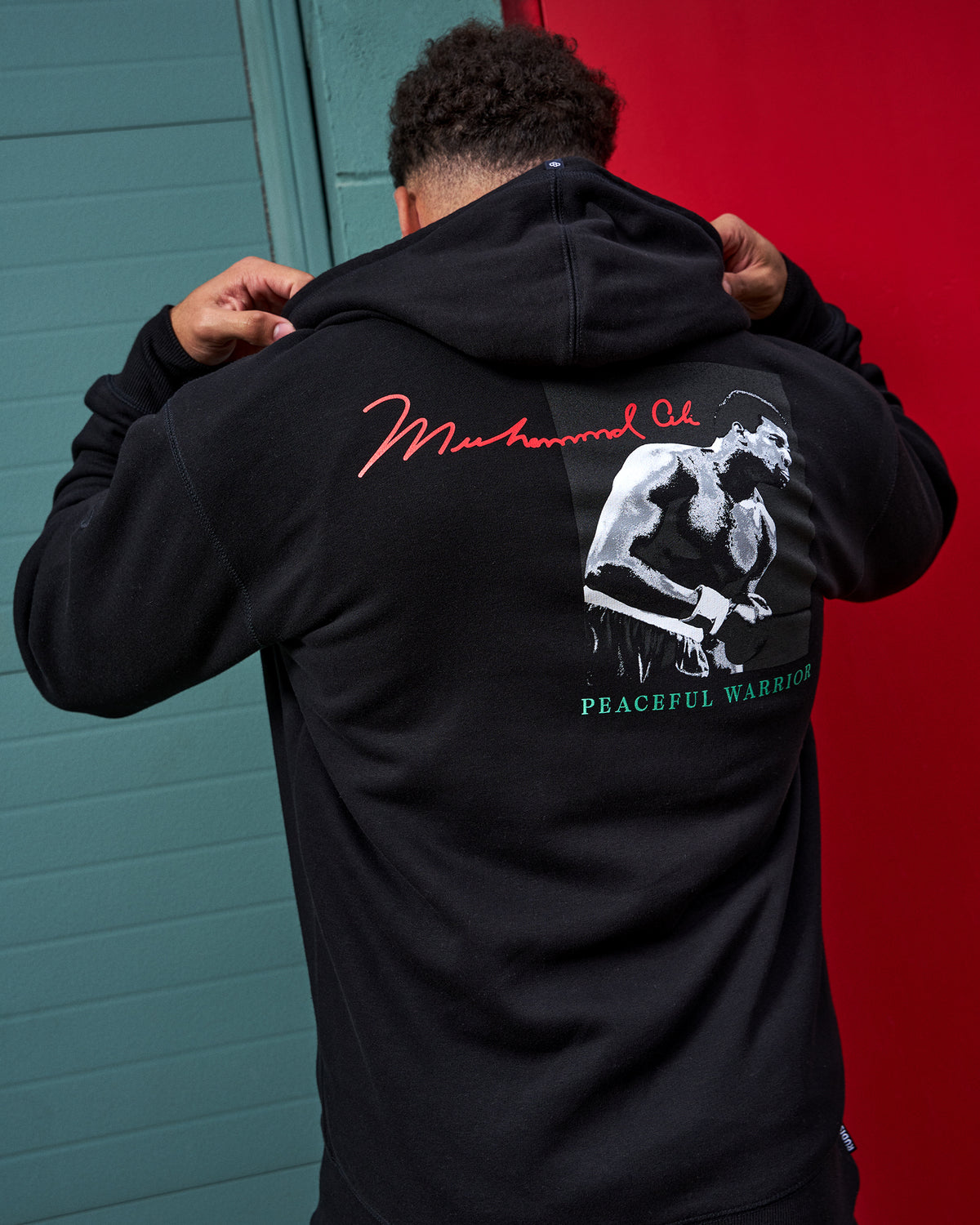 Person wearing a black hoodie with 'Muhammad Ali' design on the back against a red and blue wall.