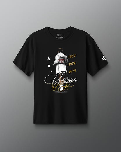 RUDIS Ali World Champion Heavyweight T-Shirt in black, featuring a Muhammad Ali-inspired championship graphic with 1964, 1974, and 1978 title years, World Champion script, and RUDIS Hex sleeve branding.