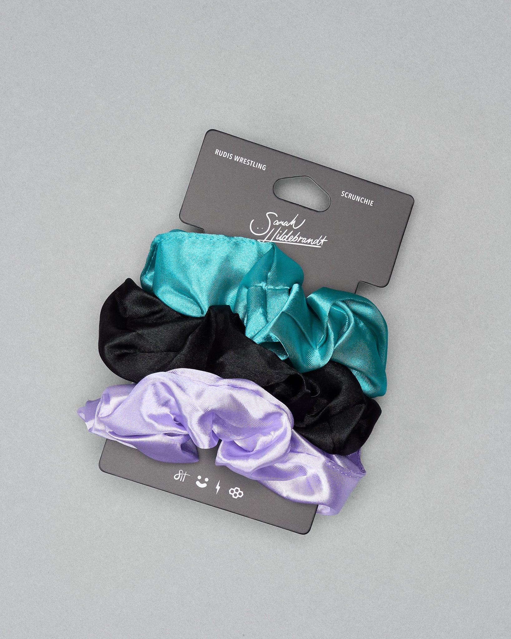 Set of three satin scrunchies (black, teal, purple) on a gray background with a brand card.