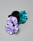 Three satin scrunchies in purple, black, and teal on a gray background