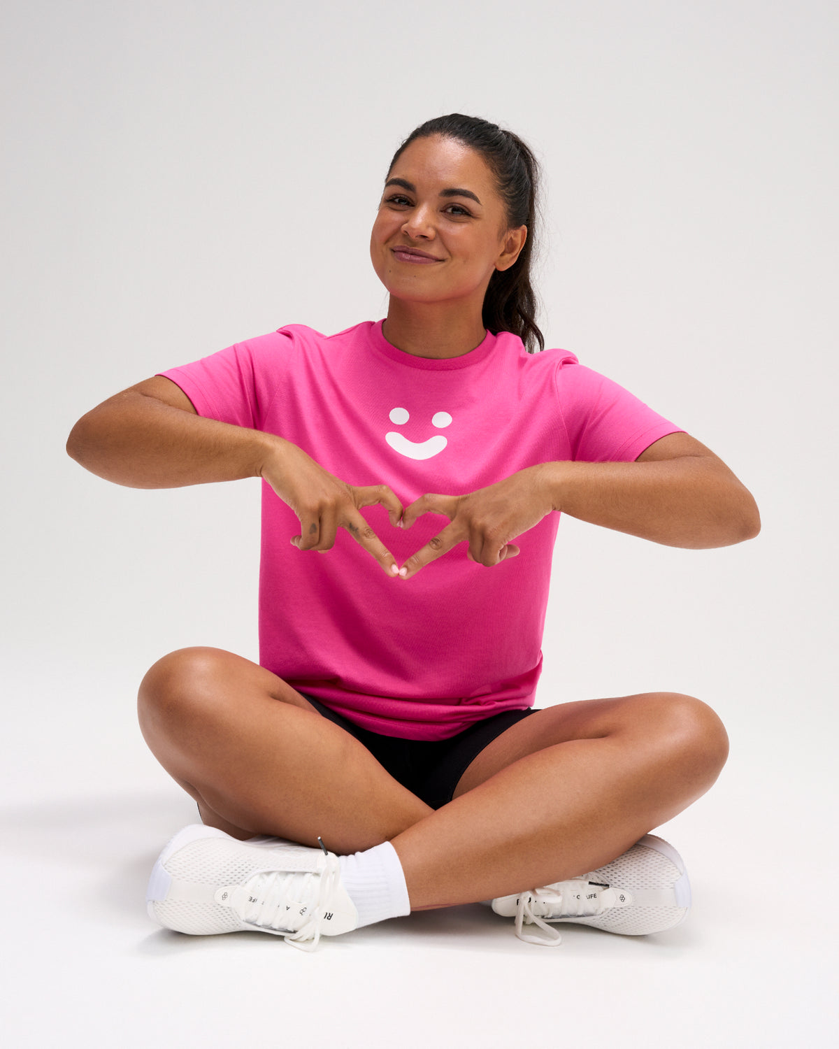 Woman wearing the pink RUDIS SH Smile Elite Super Soft T-Shirt with white smile graphic while seated in black athletic shorts and white shoes.