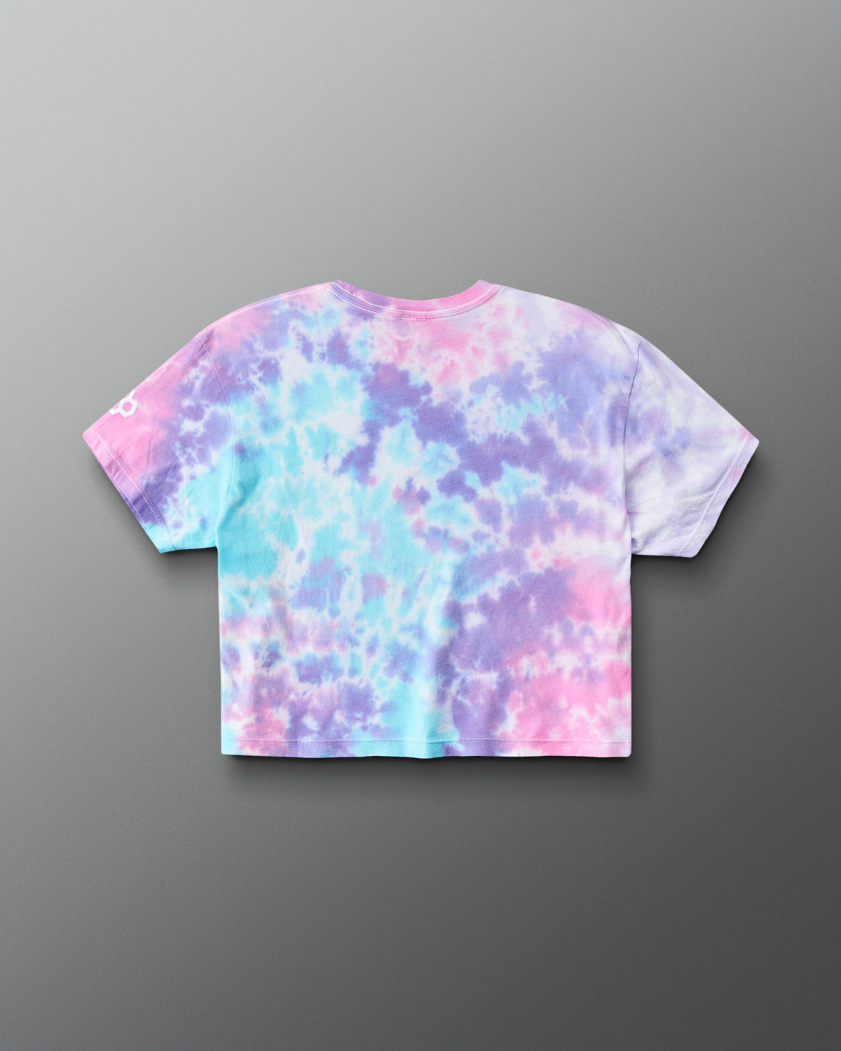 A short-sleeve crop top t-shirt with a pastel tie-dye pattern in pink, purple, and light blue, shown from the back on a gray gradient background.