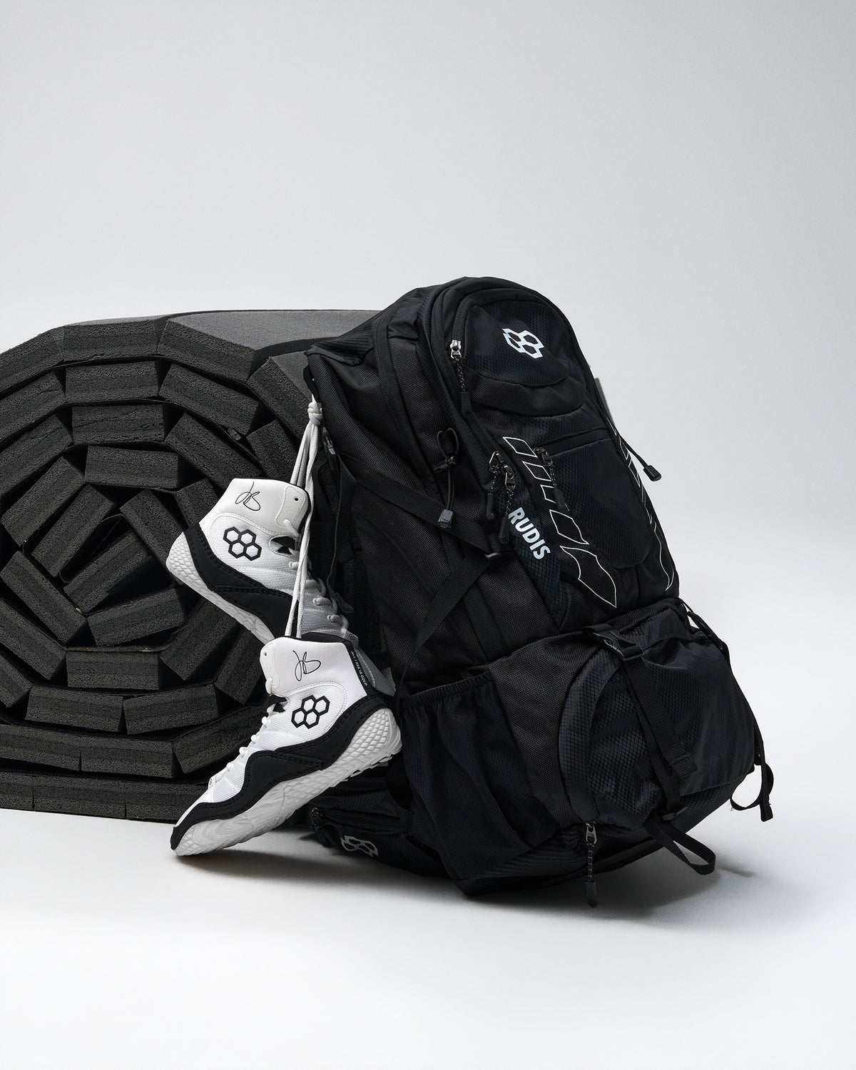 A black backpack with white logo details and a white-and-black wrestling shoes draped over its side, set against a light gray studio background with a black foam wrestling mat.