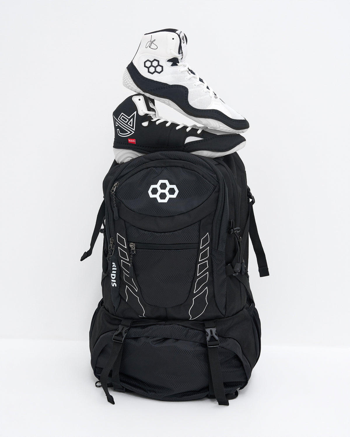 A black backpack with white graphic details and a white geometric logo on the front pocket, styled with a pair of black and white high-top wrestling shoes stacked on top, set against a plain white studio background.
