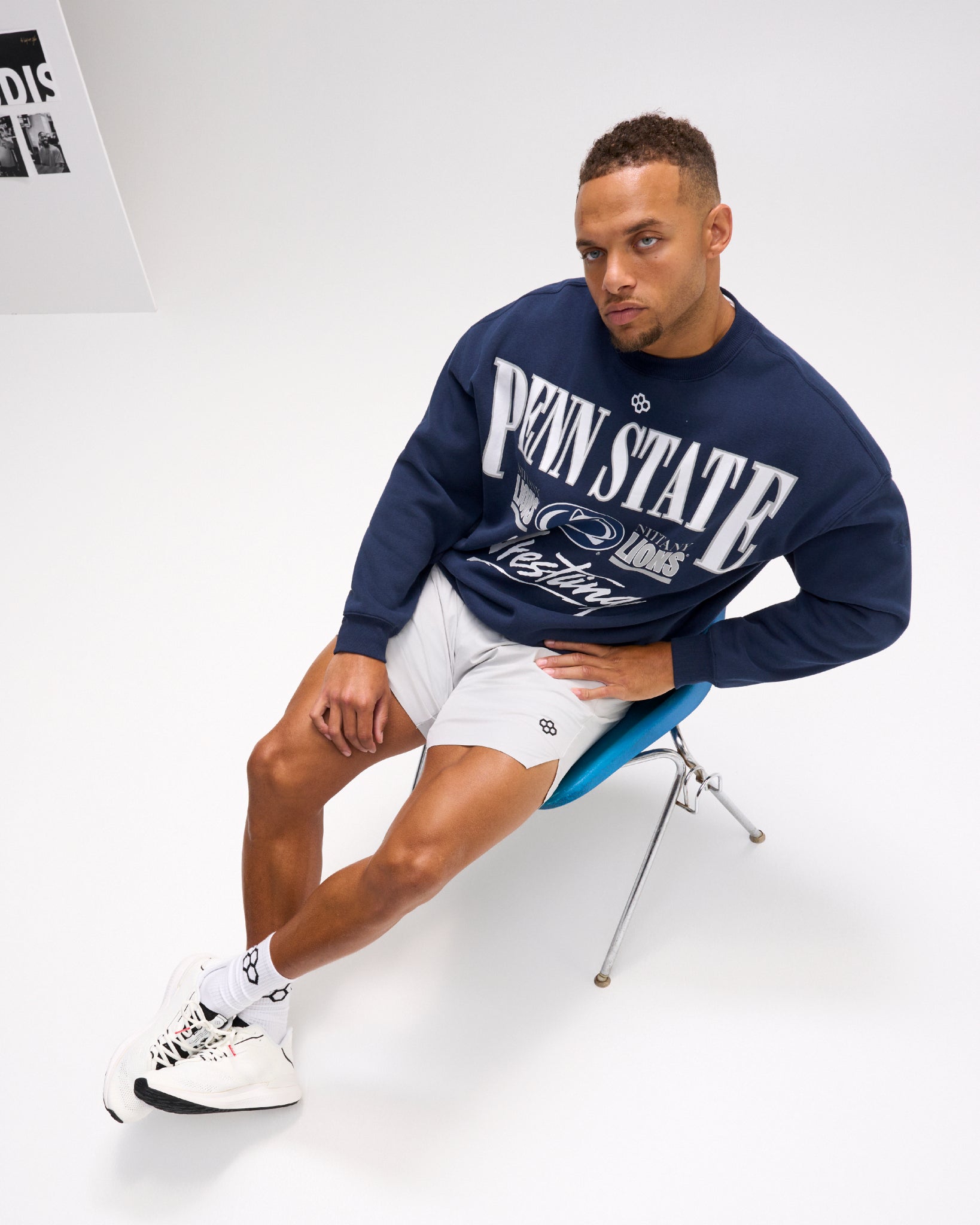 Seated male model wearing the RUDIS Penn State Map Relaxed Comfort Fleece Crewneck in Navy with bold Nittany Lions wrestling graphics, paired with white shorts.