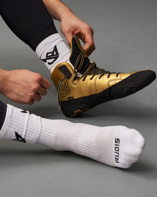 A close-up of a person putting on a vibrant gold wrestling shoe paired with white socks featuring a distinct logo showcasing an active preparation for sports