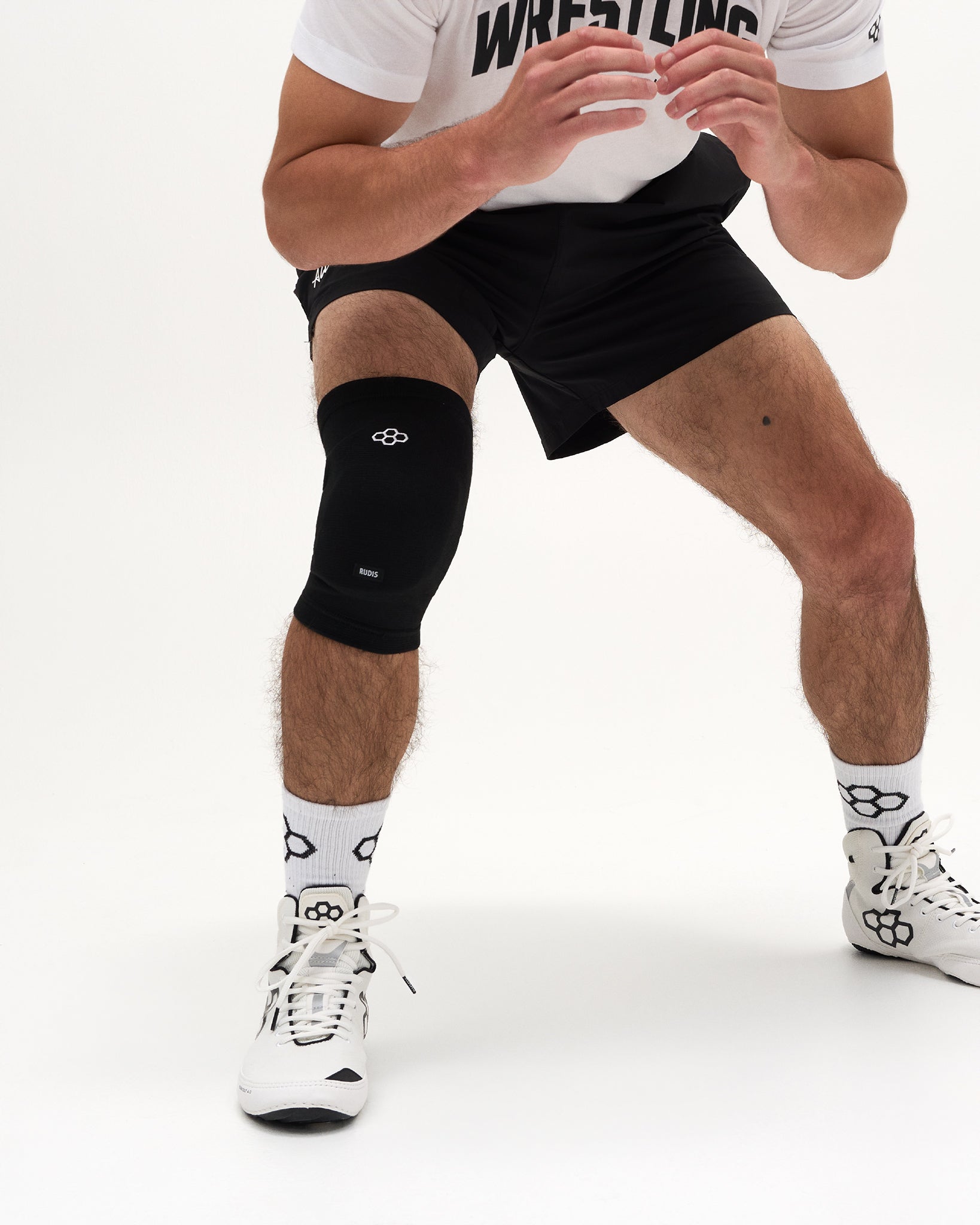 A close-up of a person's lower body wearing a black athletic knee sleeve on the right leg, black wrestling shorts, white socks, and white wrestling shoes against a clean white studio background.
