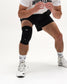 A close-up of a person's lower body wearing a black athletic knee sleeve on the right leg, black wrestling shorts, white socks, and white wrestling shoes against a clean white studio background.