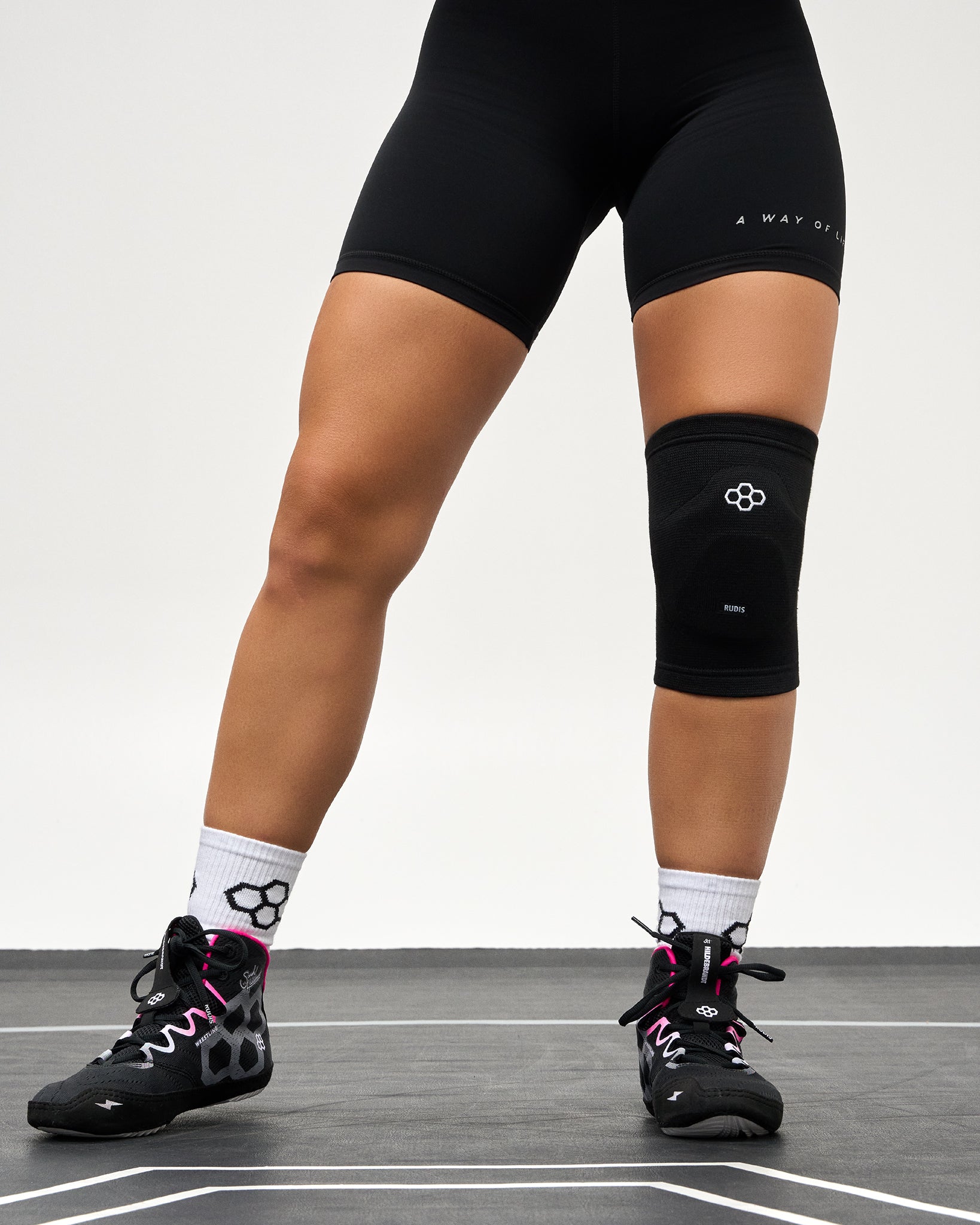 A person wearing black athletic shorts, a black knee support brace on one leg, white socks, and black running shoes with pink laces, standing on a light gray indoor court floor.