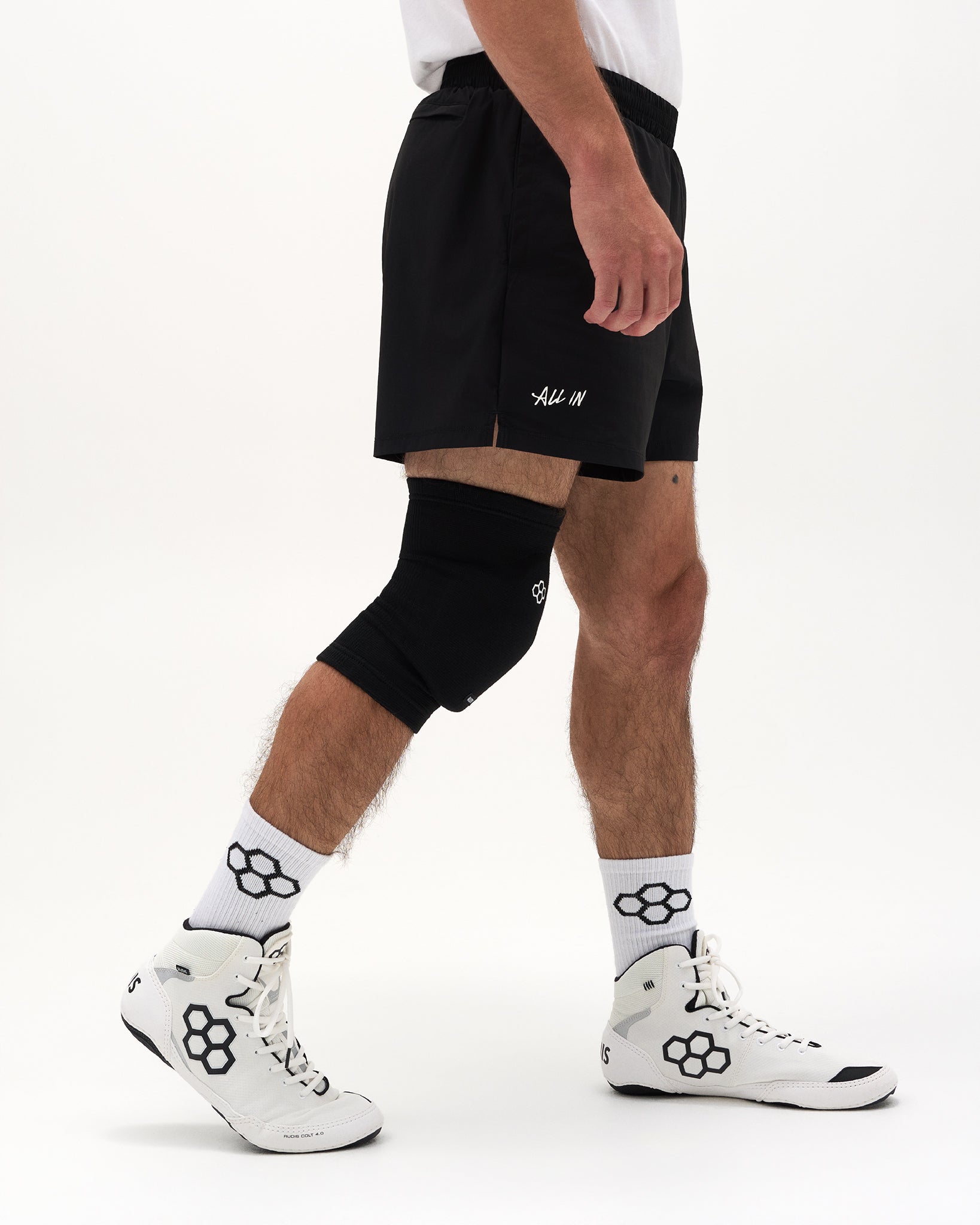 A person wearing black athletic shorts with a small white script logo, a black knee sleeve, white crew socks with a black floral pattern, and white high-top sneakers, shown against a plain light gray studio background.