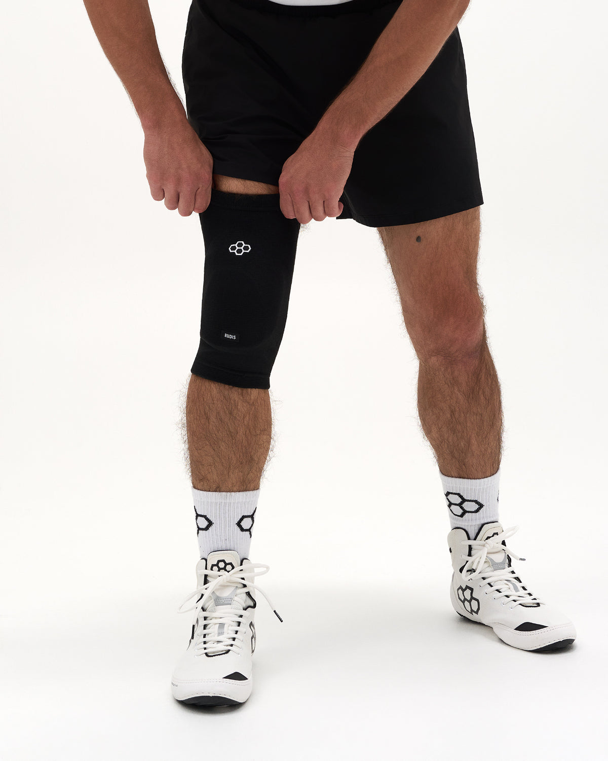 A person wearing a black knee compression sleeve on one leg, paired with black athletic shorts, white patterned socks, and white running shoes, shown against a plain white studio background.