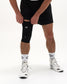 A person wearing a black knee compression sleeve on one leg, paired with black athletic shorts, white patterned socks, and white running shoes, shown against a plain white studio background.
