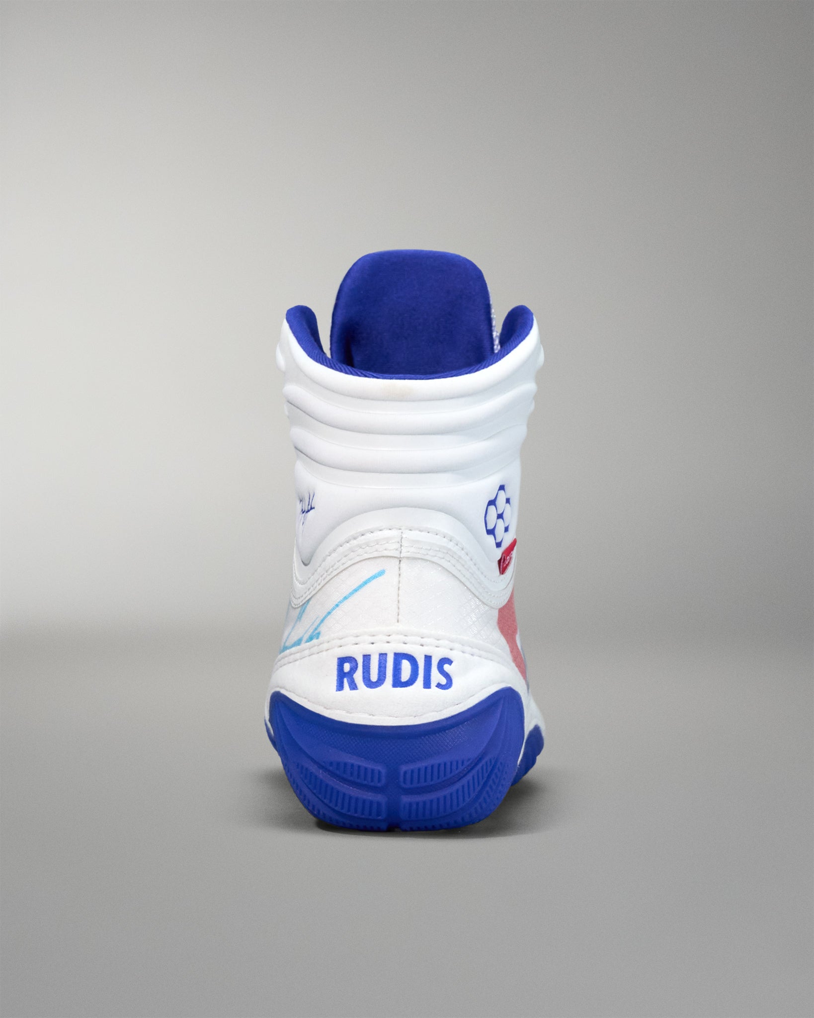 Rear view of a white RUDIS wrestling shoe with a blue sole, padded ankle collar, hexagon logo on the side, and the word ‘RUDIS’ printed in bold blue lettering on the heel.