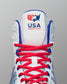 Close-up of the tongue and laces of a white RUDIS wrestling shoe featuring the USA Wrestling logo in red and blue stitching above red laces with blue accents.