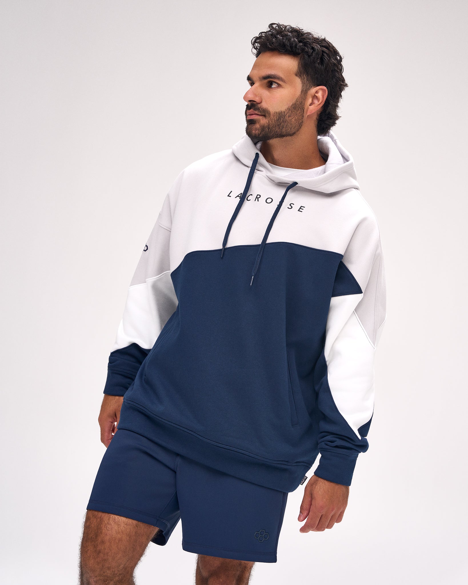 A man wearing a navy blue and white color-blocked hoodie with 'LACROSSE' printed on the chest, paired with matching navy shorts, against a light gray studio background.