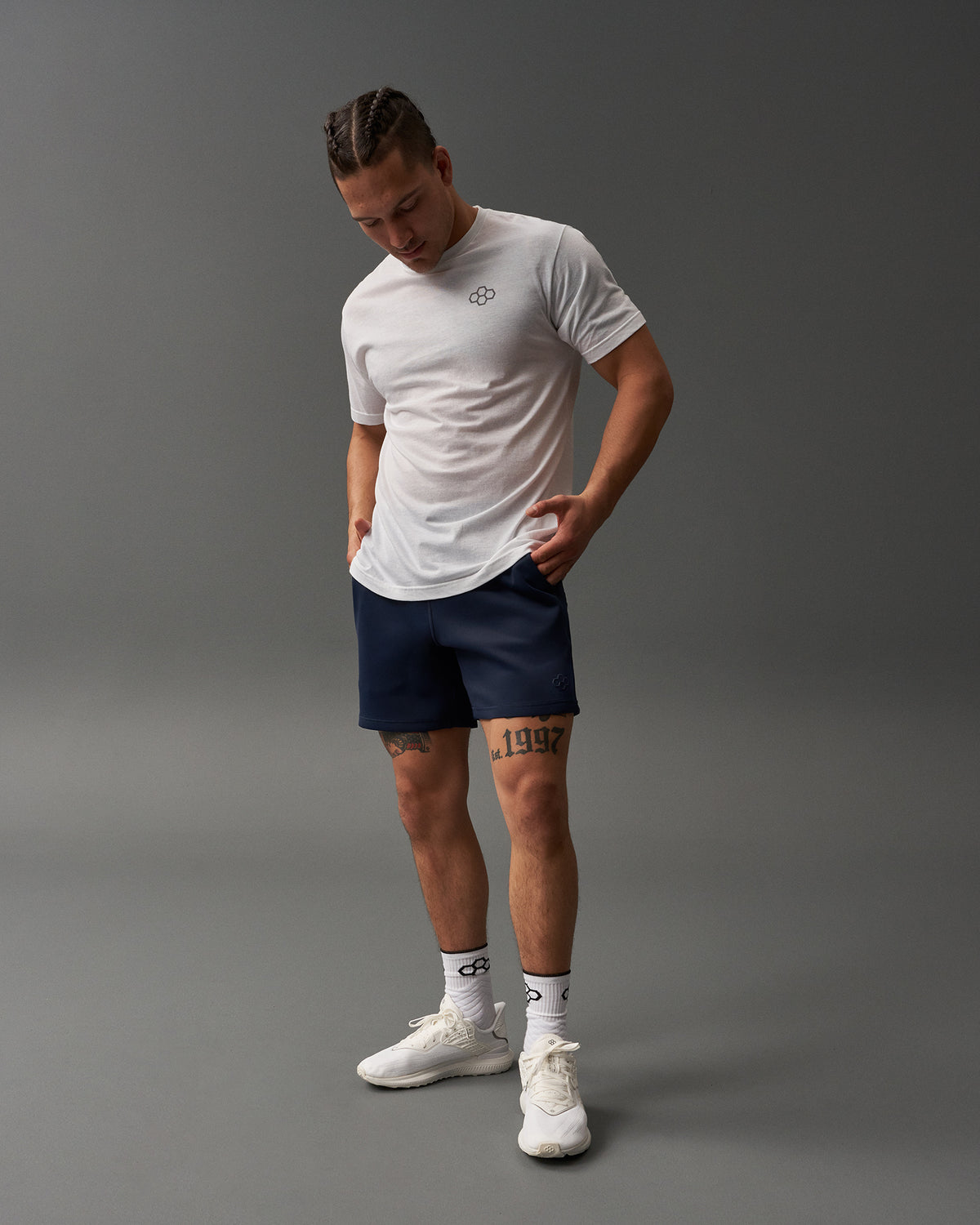 A young man with braided hair is wearing a white t-shirt and navy athletic shorts standing confidently against a muted gray background
