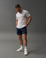 A young man with braided hair is wearing a white t-shirt and navy athletic shorts standing confidently against a muted gray background