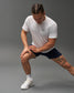 A man in a white t-shirt and navy shorts is stretching his leg highlighting a tattoo and athletic footwear against a neutral background