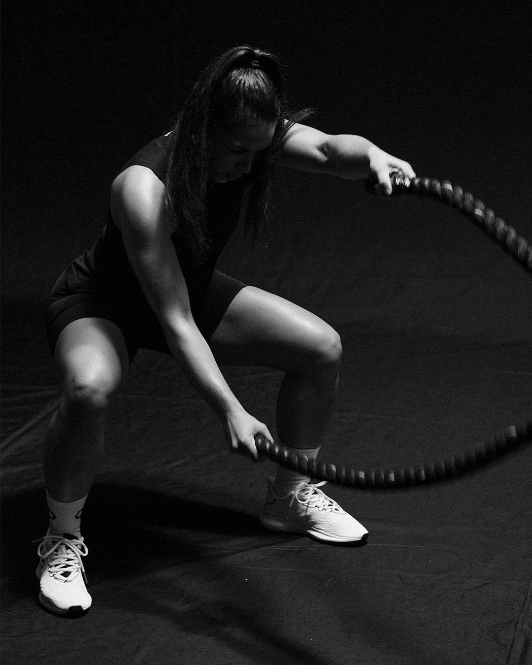 A woman training in black RUDIS athletic apparel and white sneakers while holding a heavy battle rope, emphasizing strength, conditioning, and functional fitness.