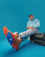 A person wearing a light blue hoodie and gray sweatpants sits next to a vintage cassette player showcasing vibrant orange and blue shoes