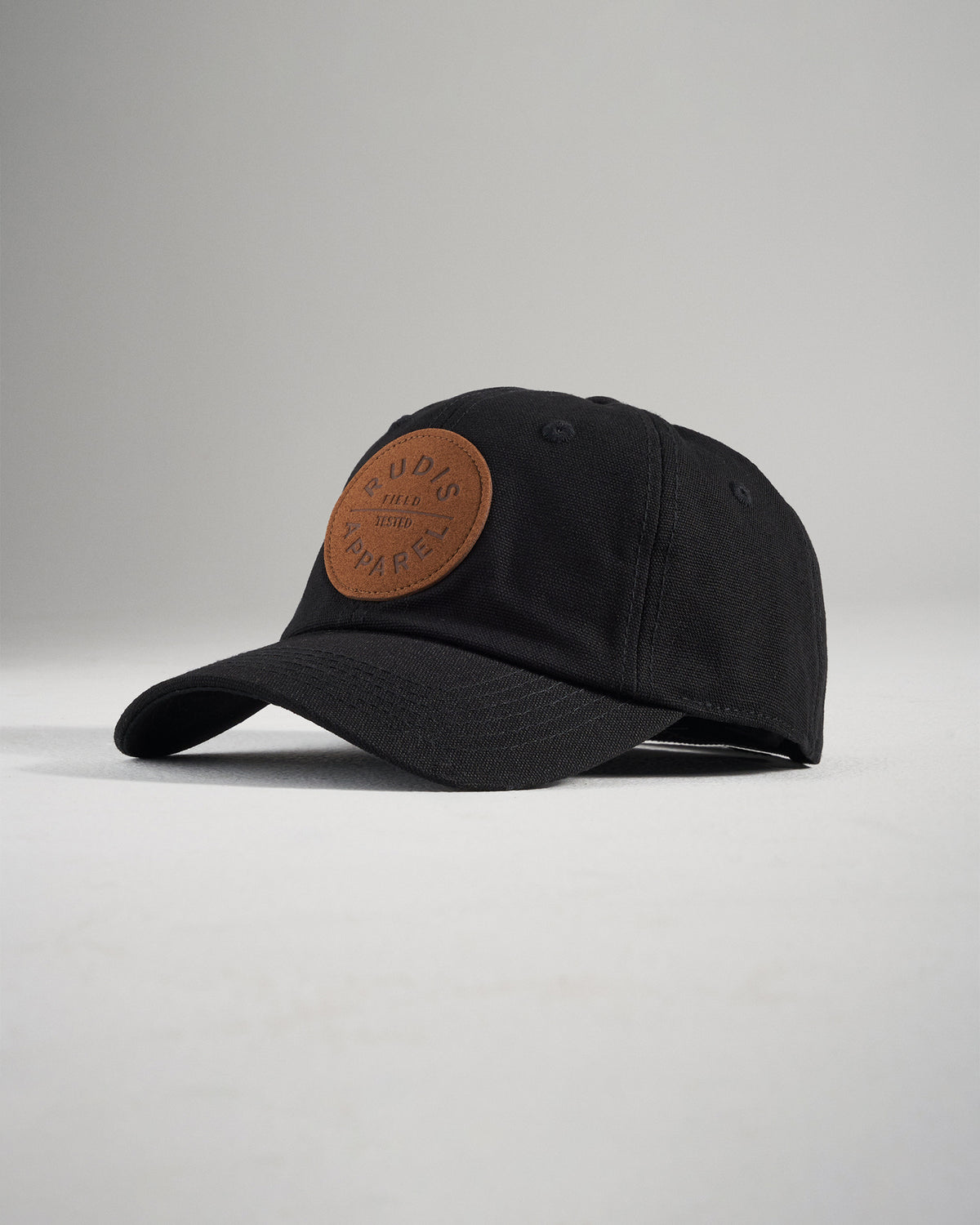 This image features a black cap with a circular leather patch labeled RUDIS APPAREL on the front set against a soft gray backdrop