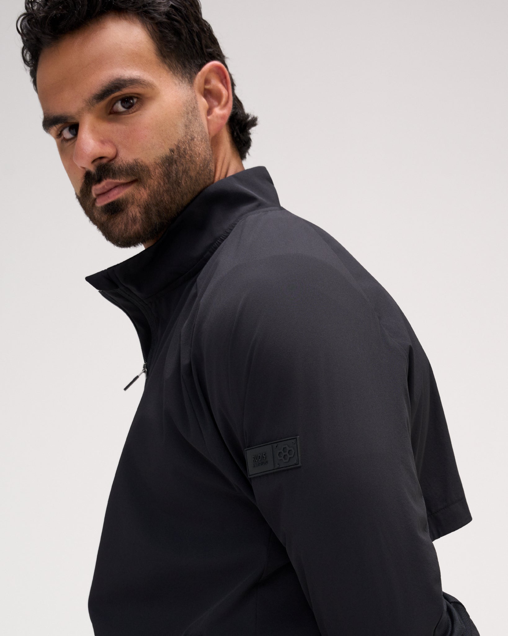 A man wearing a black zip-up athletic jacket, shown in a close-up side profile against a light gray studio background. A rectangular brand patch is visible on the upper sleeve.