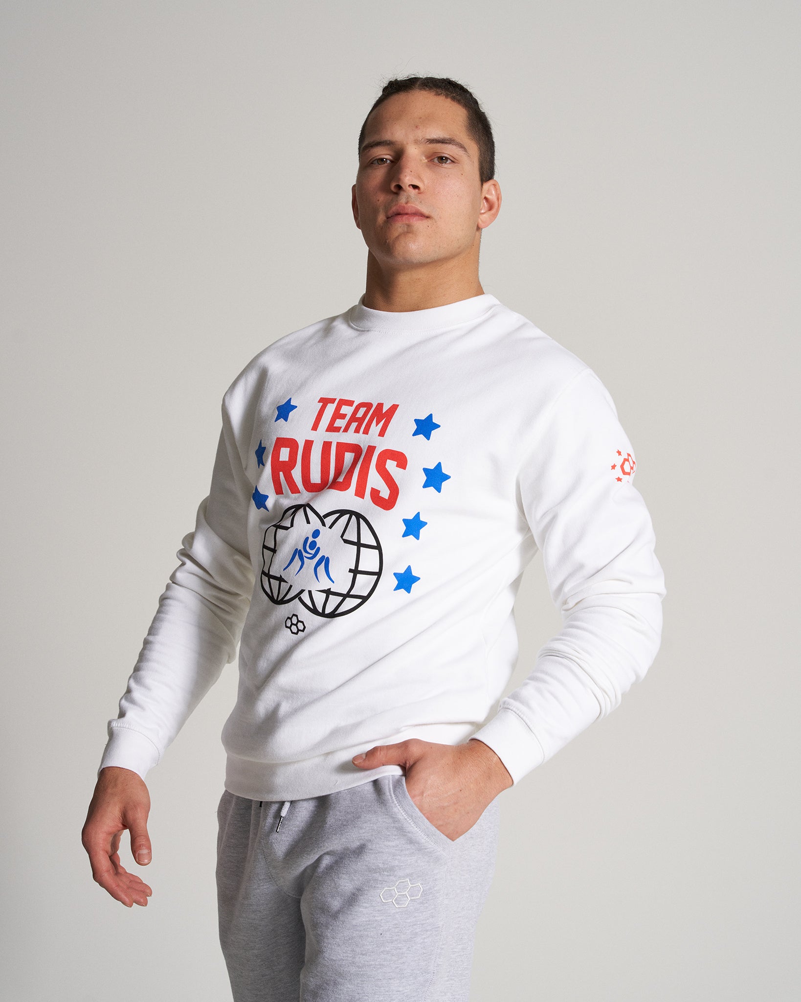 A confident athlete wearing a white sweatshirt with TEAM RUDIS printed in red complemented by gray sweatpants poses against a neutral backdrop