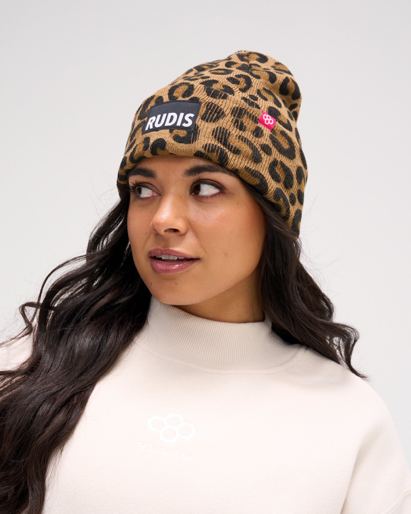 Female model wearing the RUDIS Leopard Foldover Beanie with a brown and black leopard print, ribbed foldover cuff, RUDIS logo patch and pink hex logo tag.