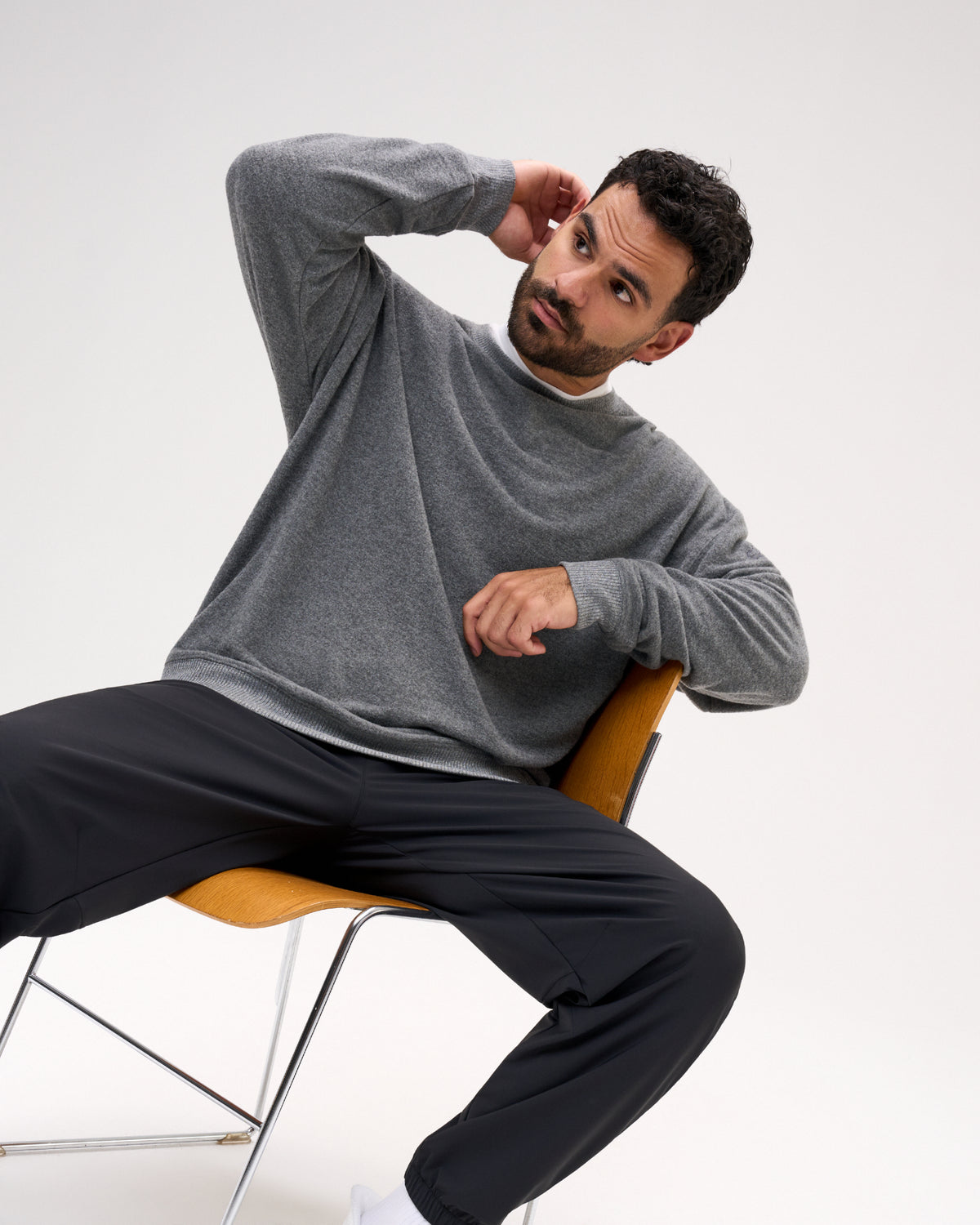 A man wearing a gray long-sleeve knit sweater and black pants sits on a modern orange chair against a plain light gray studio background.
