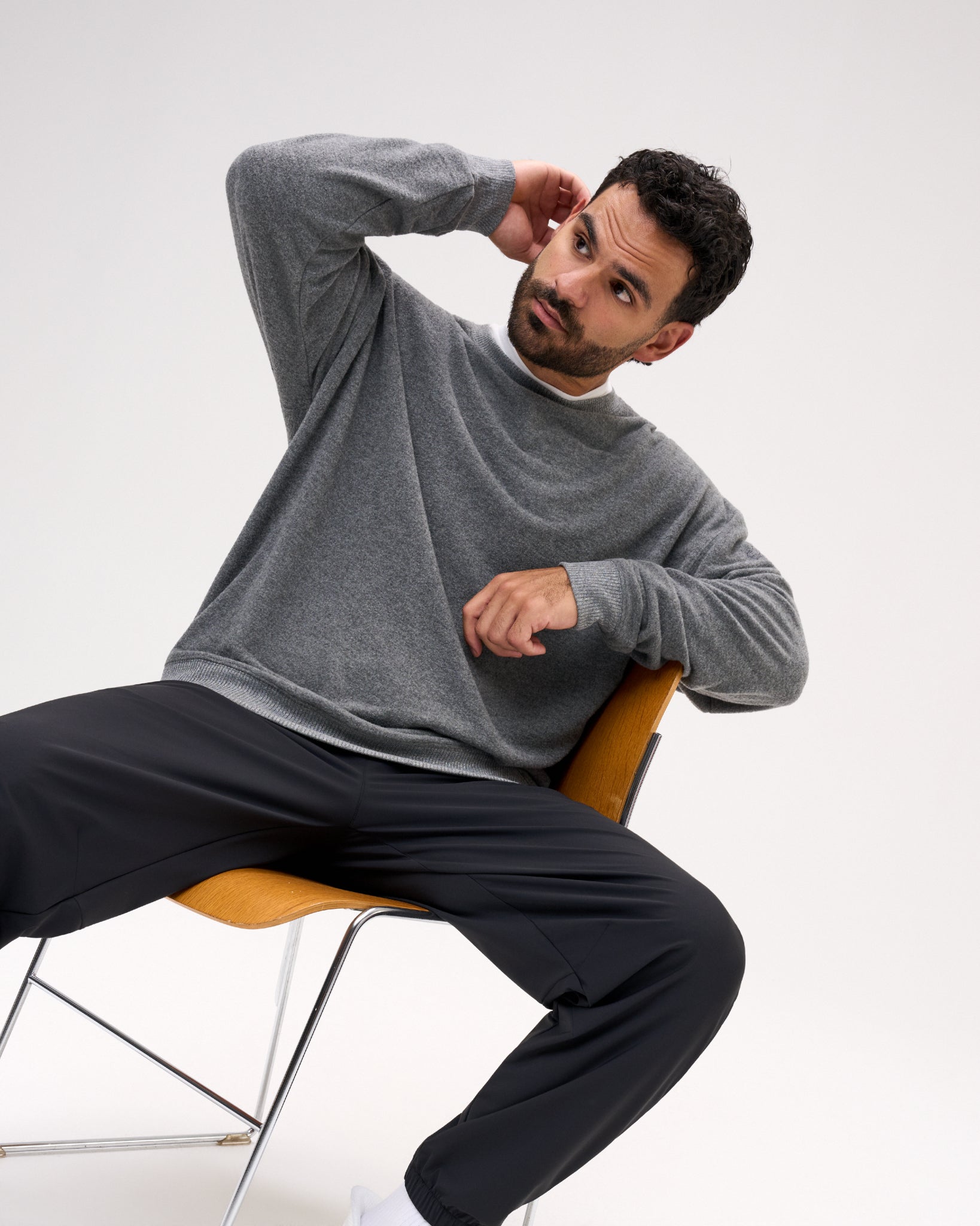 A man wearing a gray long-sleeve knit sweater and black pants sits on a modern orange chair against a plain light gray studio background.