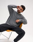 A man wearing a gray long-sleeve knit sweater and black pants sits on a modern orange chair against a plain light gray studio background.