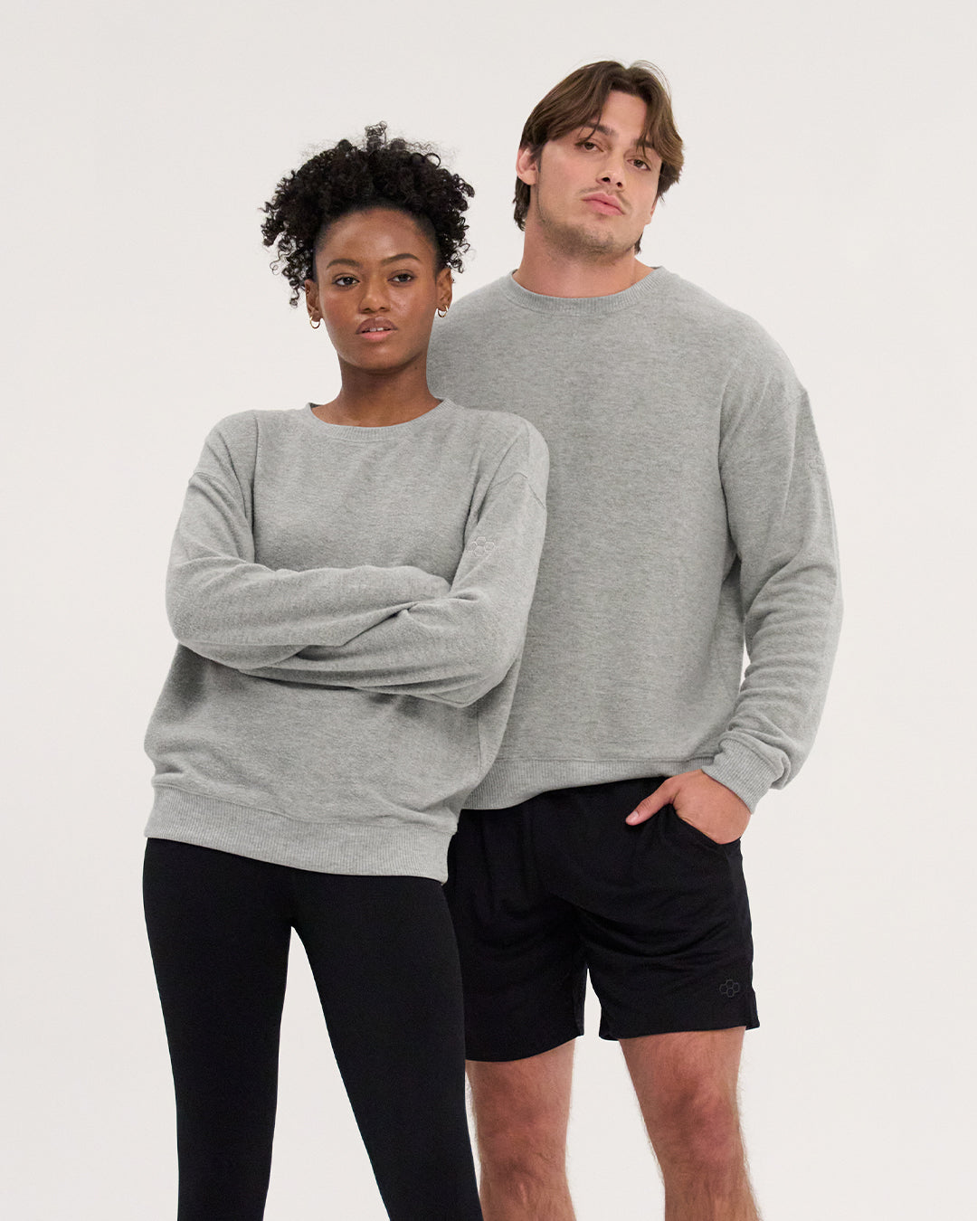 A man and a woman modeling light gray crewneck sweatshirts against a plain white studio background. The woman wears black leggings, and the man wears black shorts.