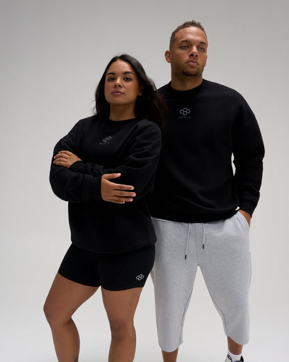 Male and female models wearing the RUDIS Hex Relaxed Comfort Fleece Crewneck in Black, featuring a tonal hex logo and relaxed fit.
 Copy alt text