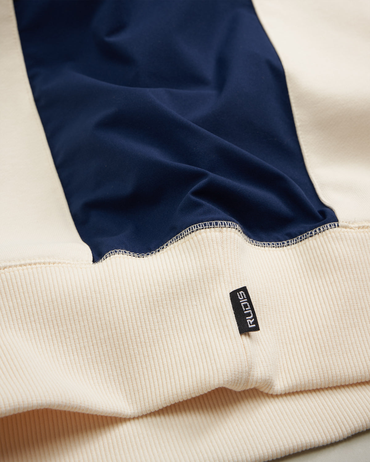 A close-up of a beige and navy color-blocked sweatshirt featuring a ribbed hem and a tag labeled RUIDIS