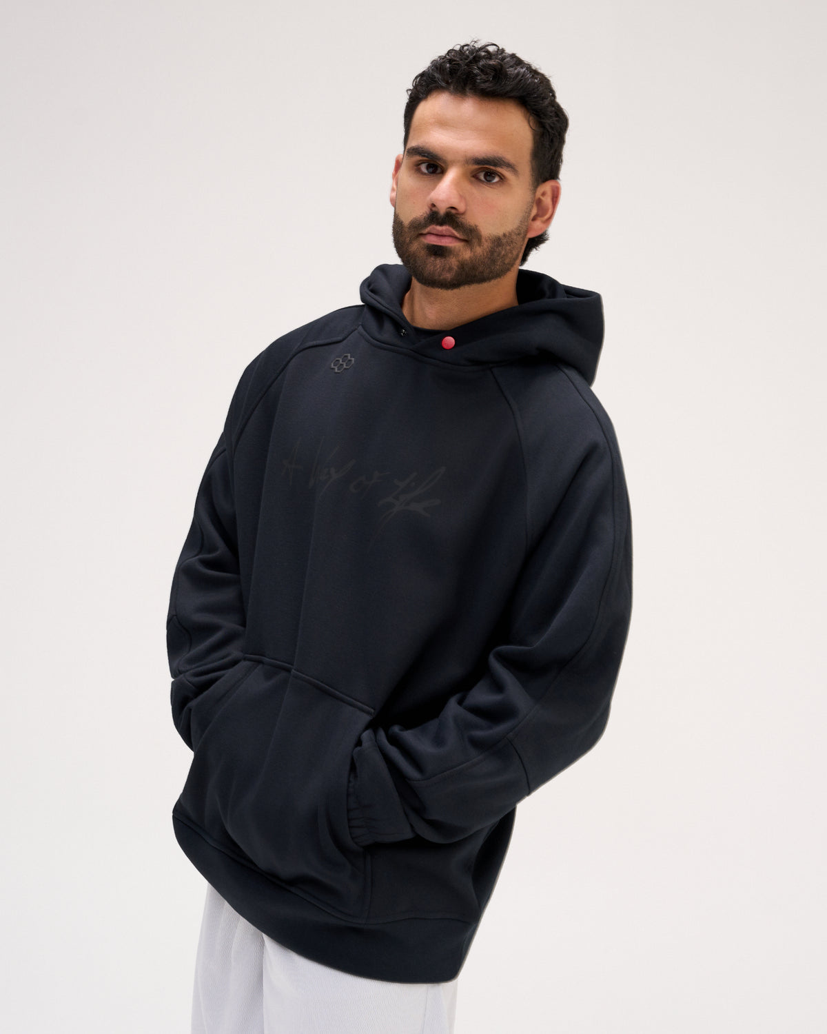 A man wearing a black pullover hoodie with a subtle tonal graphic on the chest, paired with white pants, photographed against a plain light gray studio background.