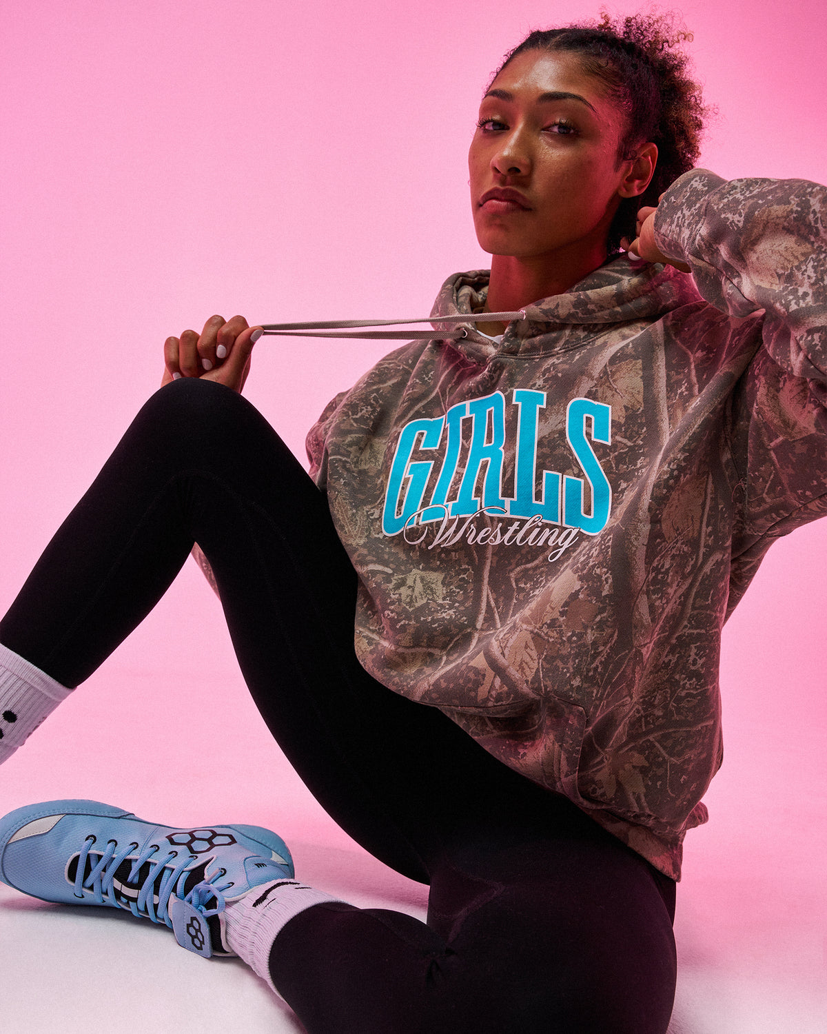 A woman sits against a pink studio background wearing a camouflage-patterned hoodie with 'GIRLS Wrestling' printed in bright blue and white across the chest, paired with black leggings and light blue sneakers.