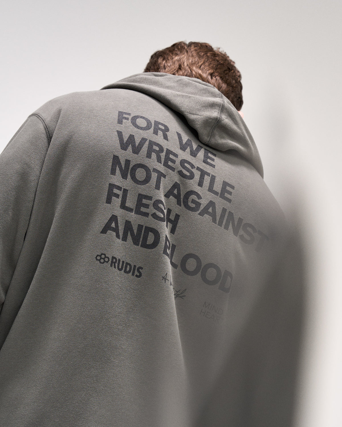 Person wearing a dark olive hoodie with 'For we wrestle not against flesh and blood' text and a brand logo on the back against a plain background