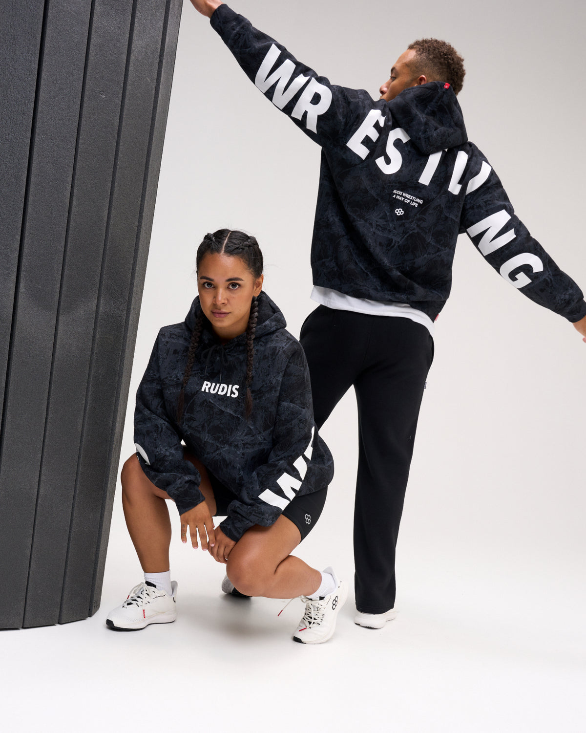 Two models posing in matching dark navy and black camouflage hoodies with white graphic text, set against a studio backdrop with a dark gray column.