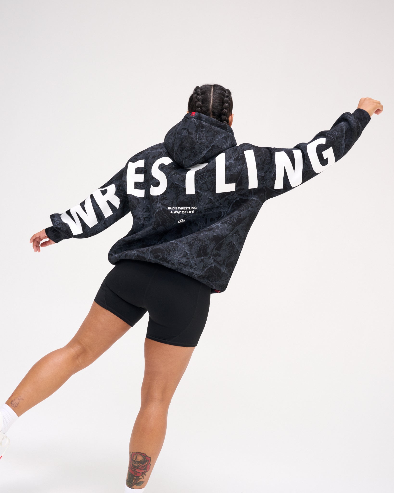 A person shown from the back wearing a dark gray and black marbled hoodie with large white 'WRESTLING' text across the back, paired with black bike shorts, against a plain white studio background.